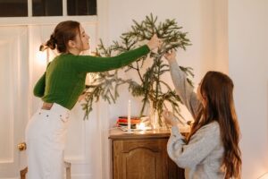 Two women decorating a small evergreen tree with lights in a cozy room.